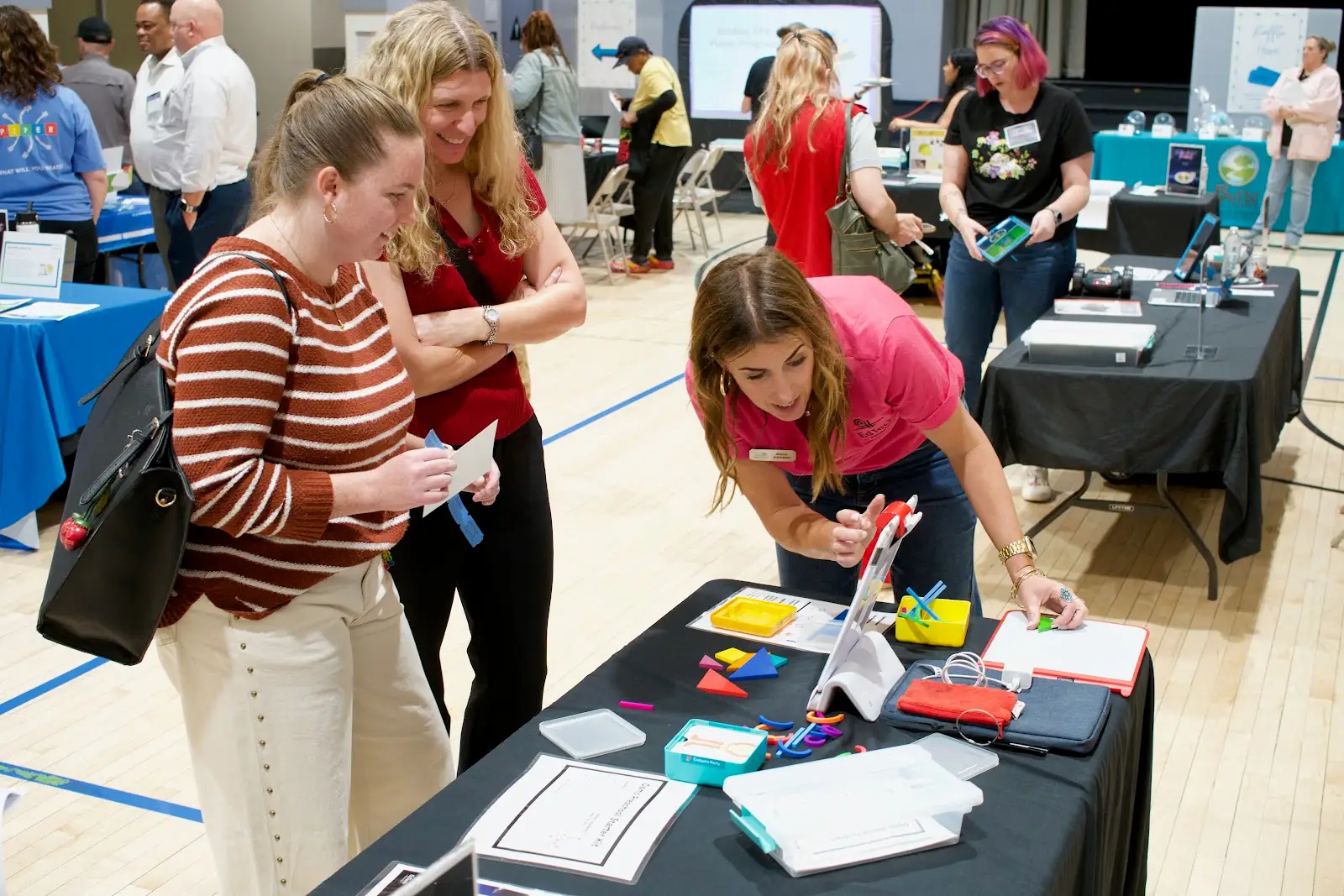 An EdTech coach shows two teachesr how to use a tablet-based tool, with many other exhibits in the background inside of a gymnasium